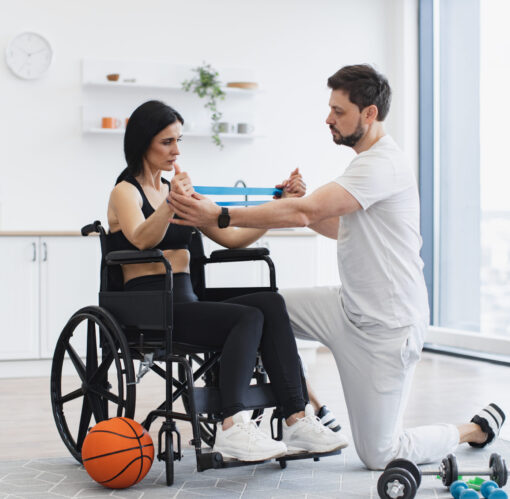 Concept of rehabilitation of disabled people. Physiotherapist or husband in sportswear helping female patient in wheelchair exercise at home. Handicapped mature woman training with stretching bands .